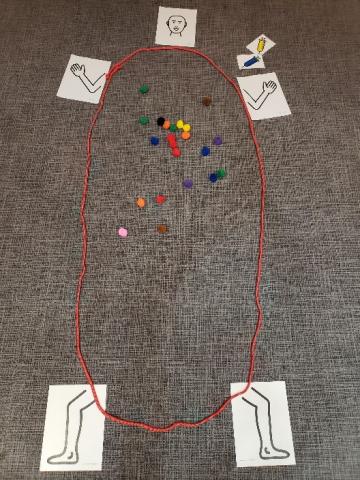 The photo shows a large circle of rope on the floor representing a human body with cards arranged to indicate the head, arms, and legs. There are many colorful pompoms inside the rope to represent germs, and pictures of blue and yellow colored syringes to represent vaccines.